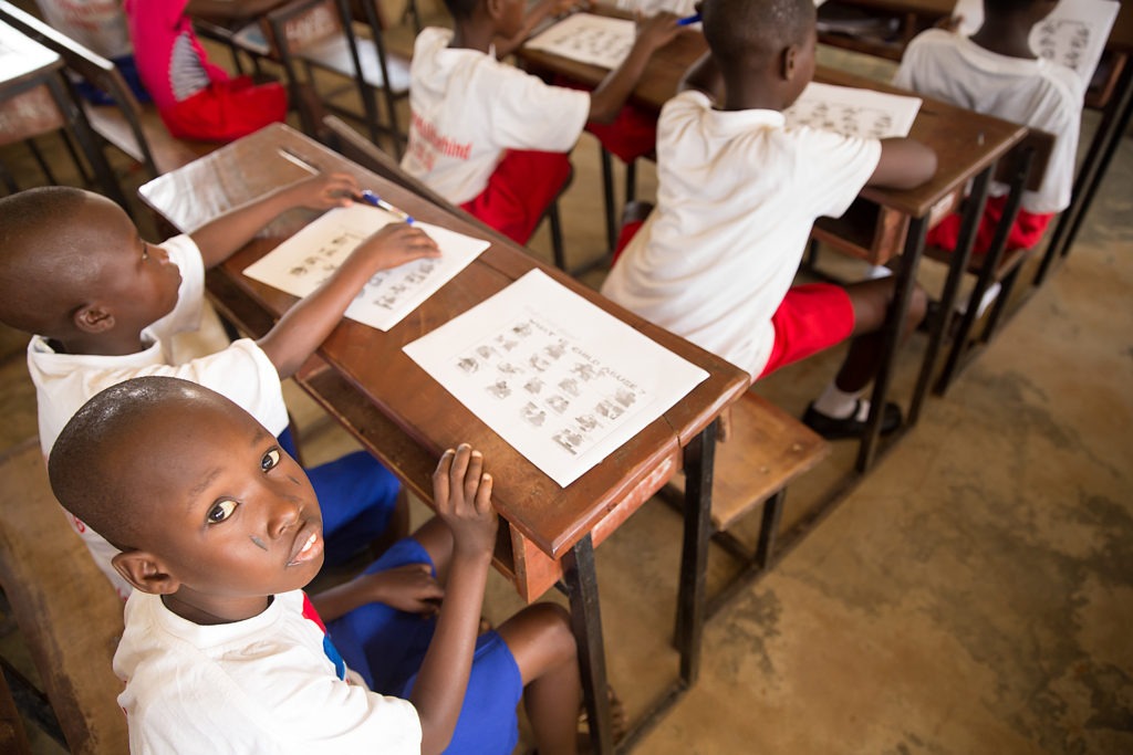 Picture of a little boy wearing a white shirt and blue shorts sitting in a classroom