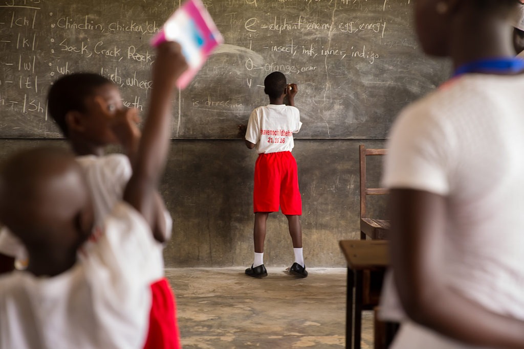 Little boy in white shirt and red shorts writing with white chalk on a chalk board